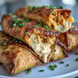 A hand holding a crispy Buffalo Chicken Dip Egg Roll, revealing a creamy, cheesy filling with green onions.